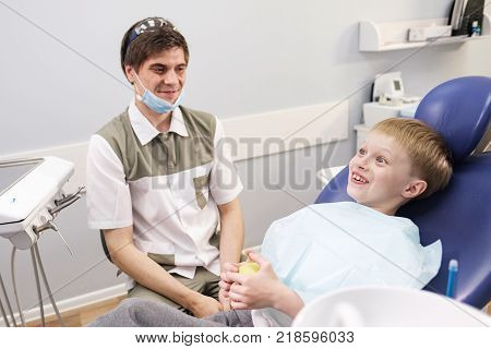 Doctor male dentist giving green apple to patient boy in dental chair