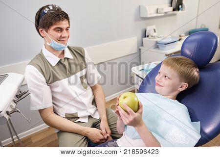 Doctor male dentist giving green apple to patient boy in dental chair