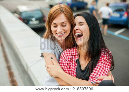 Two young teens hugging, laughting and sitting near road in the street. one brunette girl in red plaid shirt, another redhead girl wearing gray shirt and blue skirt. concept of sincere friendship