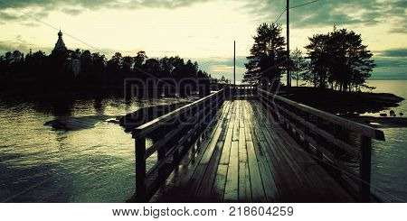 Pedestrian timber bridge on the Valaam island. Cloudy summer evening. Sunset. Rainy day. Russian north. Across the water. Valaam island Karelia Russia.