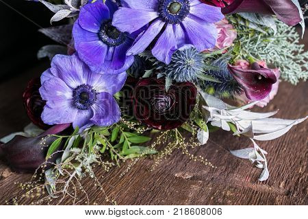 floral design, wedding, spring concept. on wooden table threre is multicolored bouquet, among beautiful flowers such as blue and lilac anemons, pink lilies and puffy battercups there are also thistles