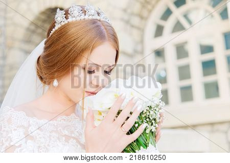 Smiling Young bride in white dress is sniffing her wedding bouquet which is carefully held by hands. The concept of florists at wedding events