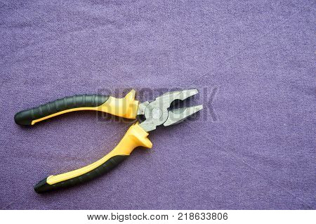 Metal iron pliers with rubberized yellow-black handles against the background of purple cloth. Bench tools.
