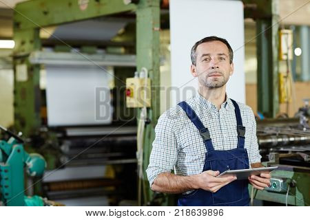 Worker of factory in overalls holding touchpad and consulting hotline for technicians