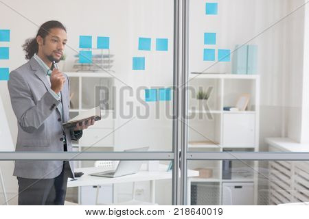 Pensive businessman with open notebook standing by transparent noteboard in office