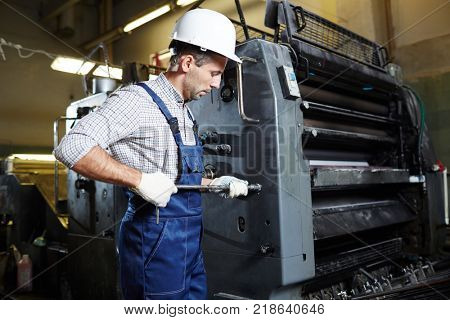 Worker with handtool standing by huge machine and checking its working condition
