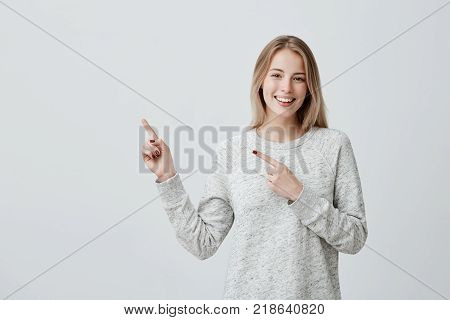 Check this out. Studio portrait of attractive positive young female with dyed blonde hair smiling cheerfully and pointing with forefingers away, indicating copy space on blank wall, having joyful happy look