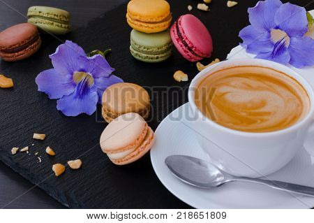 Makarons with a cup of your favorite coffee and blue flowers on a black stone background.