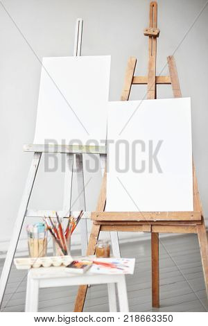 Vertical light shot of painter workplace mock up. Watercolor brushes and accessories. Two wooden easels with blank painting canvas as copy space for mock up isolated on artistic studio background.