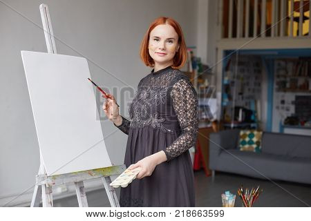 Indoor view of beautiful young Caucasian woman artist with red hair busy making drawings in spacious workshop interior with lots of paint brushes on table and mock ups around in art studio space.