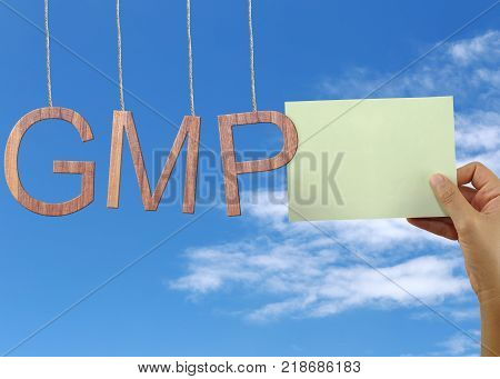 Wooden GMP text Stands for Good Manufacturing Practice hanging on rope and hand of a businessman holding a green note on blue sky background.