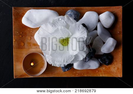 The wooden small board has a bud of a white beautiful flower, a small burning candle and a multiform pebble, for spa procedures. Close-up. View from above.