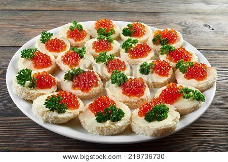 White plate on the wooden table, full of little canapees with butter, red caviar and decorated with fresh green parsley leaves. Tasty appetizer foe alcohol catering or restaurant buffet