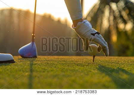 Hand asian woman putting golf ball on tee with club in golf course on evening and sunset time a for healthy sport. Lifestyle Concept