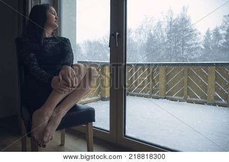 Woman relaxing in front of window while snowing - Beautiful woman wearing a sexy black dress sits on a chair with her eyes closed in front of big glass doors while outside is snowing abundantly.