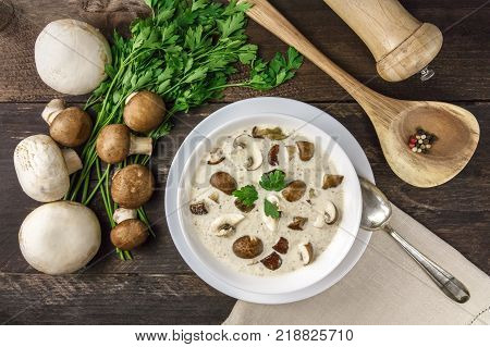 An overhead photo of a plate of a mushroom cream soup with champignons, cremini, fresh parsley, and peppercorns, on a dark rustic background texture with a place for text