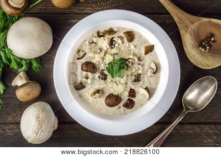 An overhead photo of a plate of a mushroom cream soup with champignons, cremini, fresh parsley, and peppercorns, on a dark rustic background texture