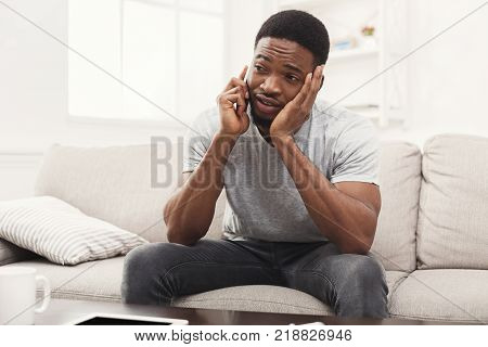 Sad young african-american man talking on smartphone emotionaly sitting on beige couch at home , copy space