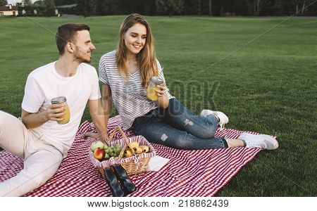 Cut view of couple whom has a small vacation and decided to go to park and sit there with some fruits and juice and just relax. Boy is looking to a girl and smiling while she is looking somewhere aside. Close up