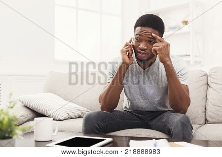 Upset young african-american man talking on smartphone emotionaly sitting on beige couch at home , copy space