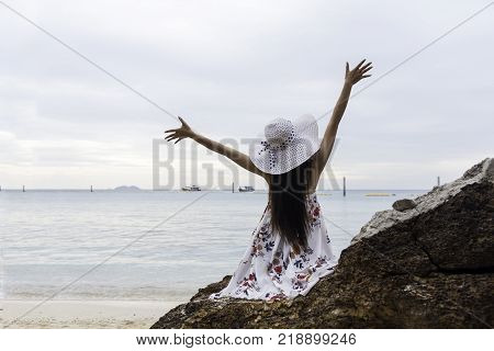Asian woman sitting at rocky beach wearing hat. White dress, casual dancing and relaxing in the solitude summer solitude in the blue sea.