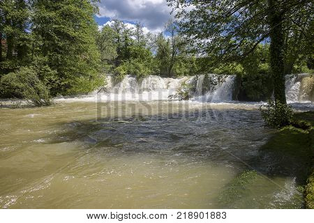Croatian Village Of Rastoke By A River Canyon With Wooden Houses In A Green Lanscape With Trees And