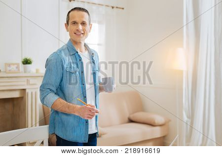 Tasty tea. Pleasant young relaxing engineer feeling good while being at home with a pencil in his hand and drinking his favorite fruit tea