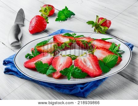 Slice of strawberry with leaf of mint and shugar on white plate on the blue linin cloth on white wooden surface.Knife o nthe second plan defocused.