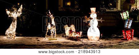 A front lawn of a home is decorated with light up Christmas decoration at night. Snowman sled angel and colorful toys boxes with sbow underneath them.