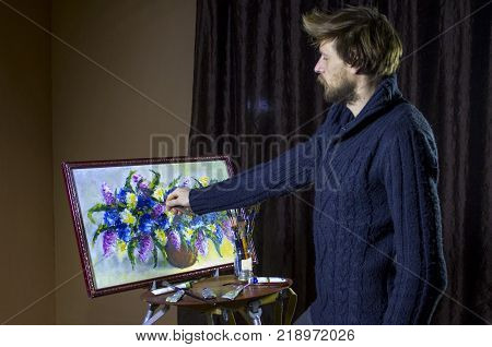 A man bearded artist in a dark sweater draws an artistic brush painting flowers still life on easer in the studio