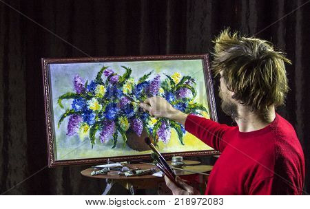 A man bearded artist in a dark sweater draws an artistic brush painting flowers still life on easer in the studio
