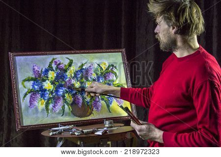 A man bearded artist in a dark sweater draws an artistic brush painting flowers still life on easer in the studio
