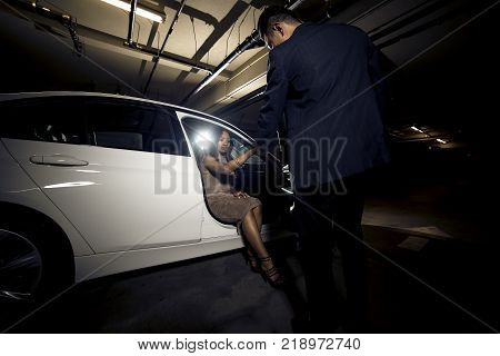 Asian male gentleman assisting his black female date out of a car. The image depicts interracial dating and manners or a vip celebrity and a valet attendant in a nightclub parking lot.