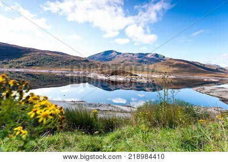 Landscape with beautiful scottish wild blooming fields and lake with mountain reflection. Panoramic view. Loch Cluanie fort William. Scotland. Uk. Vibrant colored summertime outdoors horizontal image with cloudy sky background.