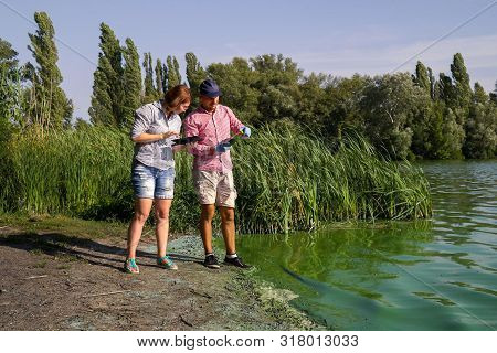 Two Ecologists Take Sample Of Green Algae On River Bank And Enter Data On Tablet