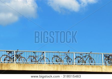 Low Angle View Of A Row Of Bicycles Parked At The Railing Of A Bridge In Front Of Blue Sky And White