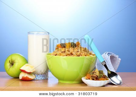 leckere Cornflakes in grüner Schale, Äpfel und Glas Milch auf Holztisch auf blauem Hintergrund