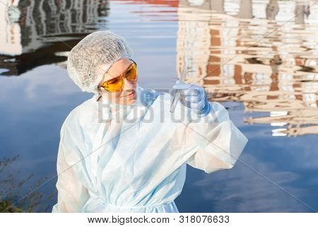 Female Epidemiologist Looks At A Test Tube With Water Against The Background Of A Water Surface With
