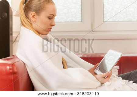 Young Woman Lying On Sofa Under Warm Blanket, Relaxing Using Tablet, Reading News And Checking Socia