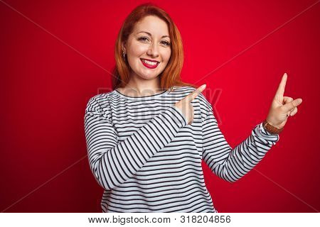Young redhead woman wearing strapes navy shirt standing over red isolated background smiling and looking at the camera pointing with two hands and fingers to the side.