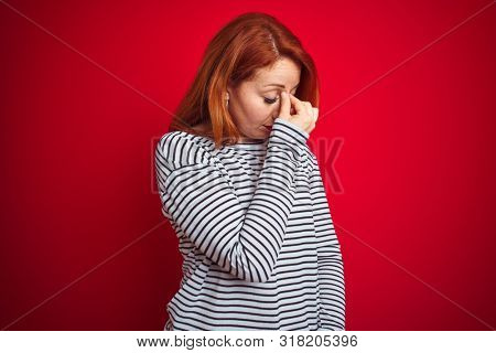 Young redhead woman wearing strapes navy shirt standing over red isolated background tired rubbing nose and eyes feeling fatigue and headache. Stress and frustration concept.