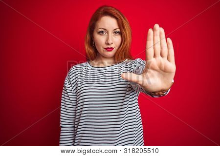 Young redhead woman wearing strapes navy shirt standing over red isolated background doing stop sing with palm of the hand. Warning expression with negative and serious gesture on the face.