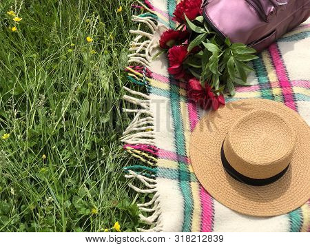 Straw Hat And Pink Backpack Full Of Red Peonies On The Checkered Blanket On The Green Grass. Copy Sp