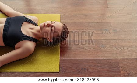 From Above Young Lady With Closed Eyes Lying In Corpse Pose On Yoga Mat On Wooden Floor In Gym