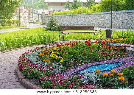 Round Flower Bed With Colorful Decorative Flowers And Plants, Garden Bench Stands On The Paving Tile