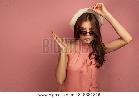 A Dark-haired Girl In A Pink Dress, Opening Her Eyes Wide, Raises A Straw Hat And Points The Lodon T
