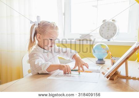 Cute Little Preschooler Girl With Two Ponytales In White Shirt Drawing At Home At The Table. Back To