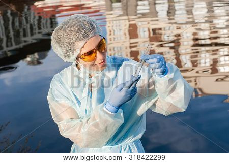 Female Epidemiologist Looks At A Test Tube With Water Against The Background Of A Water Surface With