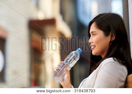 lifestyle and people concept - happy smiling young asian woman drinking water flom plastic bottle sitting on wooden city bench