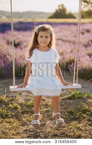 Smiling Girl On Swing Lavender Field Background. Cute Little Lady In White Dress Sitting On Teeterbo